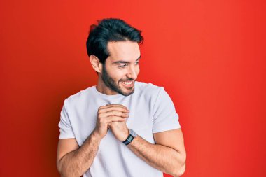 Latin asıllı genç bir adam, günlük beyaz tişört giyiyor. Gergin ve heyecanlı bir şekilde gülüyor. Elleri çenesinde yan tarafa bakıyor. 