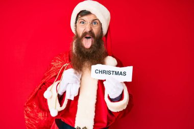 Uzun sakallı yakışıklı kızıl saçlı Noel Baba kostümü giymiş elinde Noel mesajı tutan komik bir ifadeyle dilini dışarı çıkaran genç adam.. 
