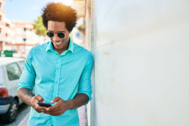 Genç, yakışıklı, Afro-Amerikan bir adam günlük giysiler giyip mutlu gülümsüyor. Yüzünde gülümsemeyle duvara yaslanıyorsun. Şehir caddesinde akıllı telefon kullanıyorsun..
