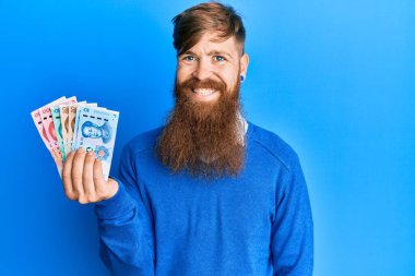 Genç İrlandalı kızıl saçlı adam Yuan Çin banknotlarını tutuyor. Pozitif ve mutlu görünüyor. Kendine güvenen bir gülümsemeyle dişlerini gösteriyor. 