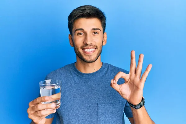 Młody Przystojny Mężczyzna Pije Szklankę Wody Robi Znak Palcami Uśmiechając — Zdjęcie stockowe