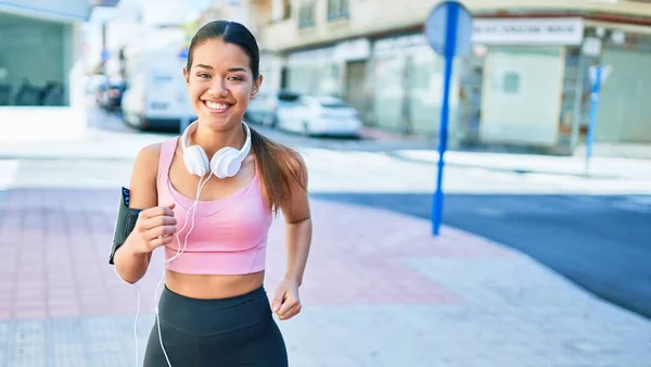 若い美しいヒスパニックスポーツの女性は ランナーの服とヘッドフォンを着て 町で走る幸せ笑顔 — ストック写真