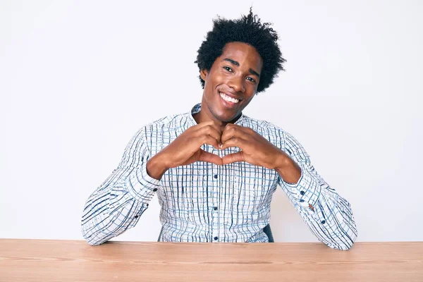 Yakışıklı Afro saçlı, sıradan kıyafetler giyen, aşık bir şekilde masasında oturan ve elleriyle kalp sembolü ve şekli gösteren Afro-Amerikan bir adam. romantik kavram. 