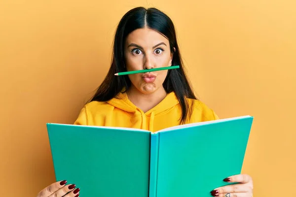 Funny hispanic woman reading a book doing crazy gesture with pencil over mouth like mustache, playful and positive on free time, lazy student