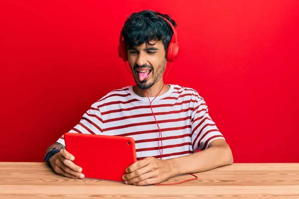 Young Hispanic Man Using Touchpad Wearing Headphones Sticking Tongue Out — Stock Photo, Image