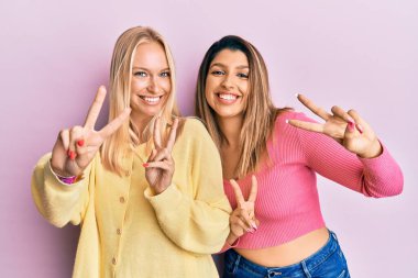 Pembe arka planda yan yana duran iki arkadaş kameraya bakıp zafer işareti yapıyor. İki numara.. 