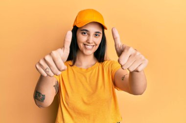 Teslimat üniforması ve şapka takan genç İspanyol kadın elleriyle olumlu bir jest yapıyor, gülümsüyor ve başarı için seviniyor. kazanan jest. 