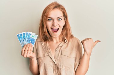 Kafkasyalı genç bir kadın, elinde 50 Polonyalı zloti banknotla baş parmağını yana uzatıyor ve ağzı açık gülümsüyor. 