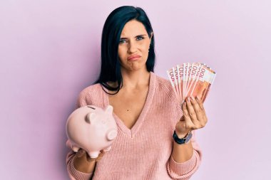 Elinde Norveç Krona banknotları tutan beyaz bir kadın ve domuz kumbarası kuşkucu ve sinirli, sorun yüzünden üzgün. negatif kişi. 