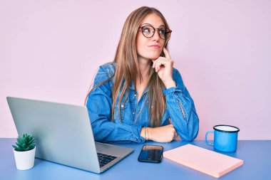 Young blonde girl working at the office drinking a cup of coffee serious face thinking about question with hand on chin, thoughtful about confusing idea 