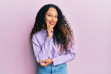 Gündelik giysiler giyen genç Latin kadın kameraya kollarını kavuşturup çenesine dokunarak gülümsüyor. Olumlu düşünmek. 