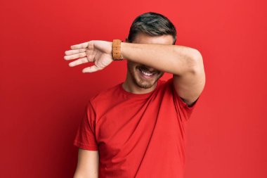 Yakışıklı, beyaz bir adam, günlük kırmızı tişört giyiyor, gözleri kollarıyla kaplıyor, neşeli ve komik gülümsüyor. kör kavram. 