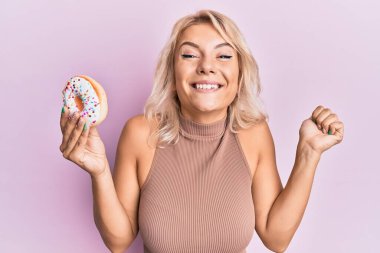 Genç sarışın kız elinde donut tutarak gururla haykırıyor zaferi kutluyor ve başarıyı kollarını kaldırarak kutluyor. 