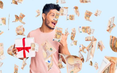 Yakışıklı Latin Amerikalı genç adam elinde hediyeyle başparmağını kaldırıyor. Gülümsüyor, ağzı açık.