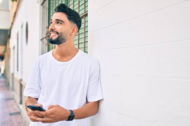 Genç Arap adam şehirde akıllı telefon kullanarak mutlu gülümsüyor..