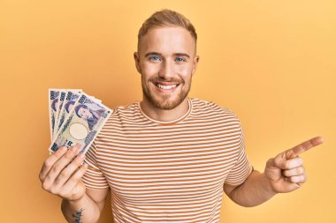 Elinde Japon Yen banknotları tutan genç beyaz adam gülümsüyor mutlu bir şekilde elini ve parmağını yana doğru gösteriyor. 