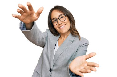 Genç esmer kadın iş kıyafetleri içinde kameraya bakıyor ve sarılmak için kollarını açmış gülümsüyor. Mutluluğu kucaklayan neşeli bir ifade.. 