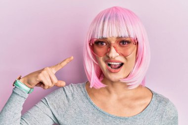 Pembe peruk ve kalp gözlüğü takan beyaz bir kadın gülümsüyor. 