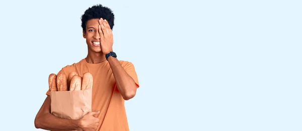若いアフリカ系アメリカ人の男性が手で片目を覆うパンで紙袋を持っています 顔に自信を持って笑顔と驚きの感情 — ストック写真