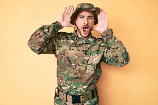 Young caucasian man wearing camouflage army uniform smiling cheerful playing peek a boo with hands showing face. surprised and exited