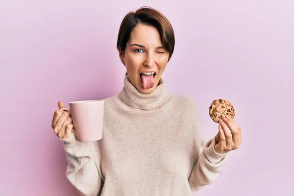 若いですブルネット女性とともに短い髪飲むAカップのコーヒーとクッキーステッキング舌アウト幸せとともに面白い式 — ストック写真