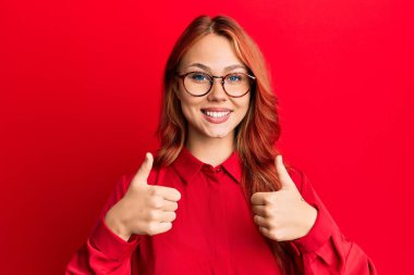 Young beautiful redhead woman wearing casual clothes and glasses over red background success sign doing positive gesture with hand, thumbs up smiling and happy. cheerful expression and winner gesture. 