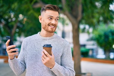 Genç beyaz adam akıllı telefon kullanıyor. Parkta kahve içiyor..