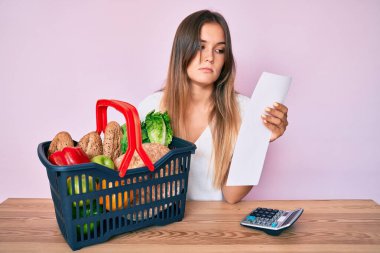 Beautiful caucasian woman holding supermarket basket and groceries list relaxed with serious expression on face. simple and natural looking at the camera.  clipart