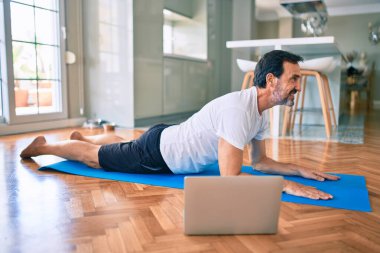 Orta yaşlı, sakallı ve esnek bir adam evde egzersiz yapıyor bilgisayardaki spor videolarına bakıyor.