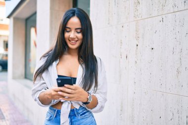 Genç İspanyol kız şehirde akıllı telefon kullanarak mutlu bir şekilde gülümsüyor..