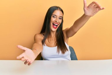 Latin asıllı genç bir kadın, gündelik giysiler içinde masaya oturmuş kameraya bakıyor ve sarılmak için kollarını açmış gülümsüyor. Mutluluğu kucaklayan neşeli bir ifade.. 