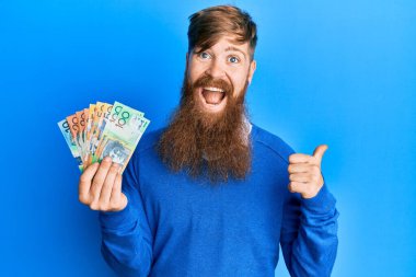 Genç İrlandalı kızıl saçlı adam Avustralya doları tutuyor başparmağını yukarı kaldırıyor ve ağzı açık gülümsüyor. 