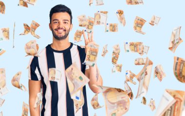 Çizgili tişört giyen genç, yakışıklı, kendinden emin ve mutlu gülümserken üç numaralı parmaklarıyla onu gösteriyor ve işaret ediyor..