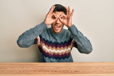 Genç, yakışıklı bir adam, sıradan bir süveter giymiş masanın üzerinde oturmuş dürbün gibi jest yapıyor, dili dışarı çıkarıyor, gözleri parmaklarından bakıyor. çılgın ifade. 