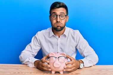 Yakışıklı İspanyol bir adam domuz kumbarası tutuyor. Gözlüklü, komik suratlı, şişen yanakları olan. Ağzı havayla şişer, havayı yakalar.. 