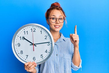 Elinde koca bir saat tutan genç kızıl saçlı bir kadın bir fikir ya da soruyla gülümsüyor ve mutlu bir yüzle işaret ediyor. 