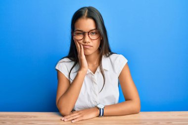 Gündelik kıyafetler giyen güzel İspanyol kadın masaya oturmuş, kollarını kavuşturup depresyondan sıkılmış ve yorgun göründüğünü düşünüyor.. 