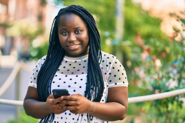 Genç Afrikalı Amerikalı kadın şehirde akıllı telefon kullanarak mutlu bir şekilde gülümsüyor..