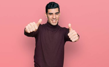 Balıkçı yaka kazak giyen yakışıklı bir adam. Elle olumlu jest yapmayı onaylıyor. Gülümsüyor ve başarı için seviniyor. kazanan jest. 