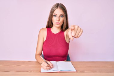 Masaya oturmuş, kamerayı işaret eden bir not defteri yazan beyaz bir kız. Kendine güvenen, ciddi görünen bir jest. 