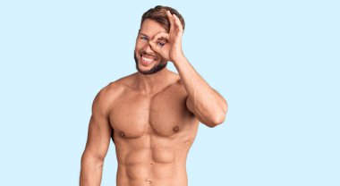 Genç beyaz adam üstsüz duruyor. El hareketleriyle gülümsüyor. Gözleri mutlu yüzüyle parmaklarının arasından bakıyor.. 