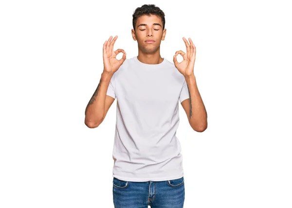 カジュアルな白いTシャツを着た若いハンサムなアフリカ系アメリカ人男性は 指で瞑想のジェスチャーをして目を閉じてリラックスして笑顔 ヨガのコンセプト — ストック写真