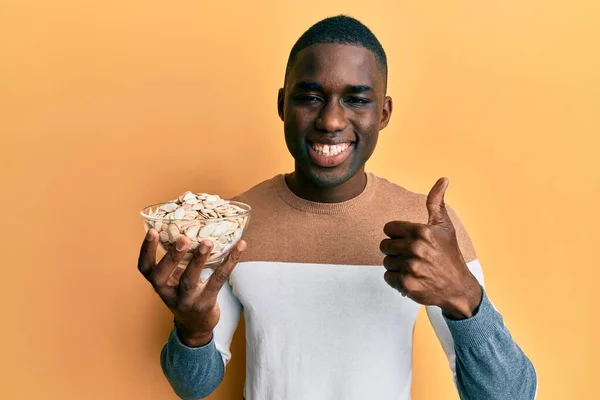 stock image Young african american man holding bowl with pumpkin seeds smiling happy and positive, thumb up doing excellent and approval sign 