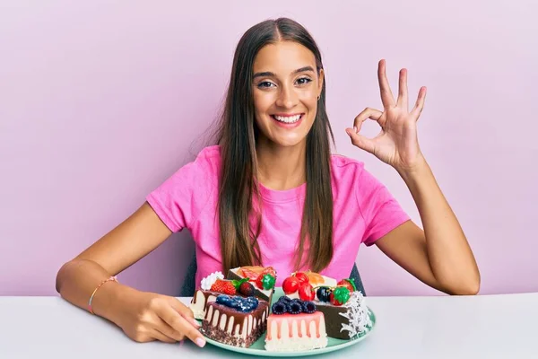 若いですヒスパニック系の女性食べるチーズケーキ上のテーブルの上に座って指でOkのサインを行います 笑顔優しいジェスチャー優れたシンボル — ストック写真