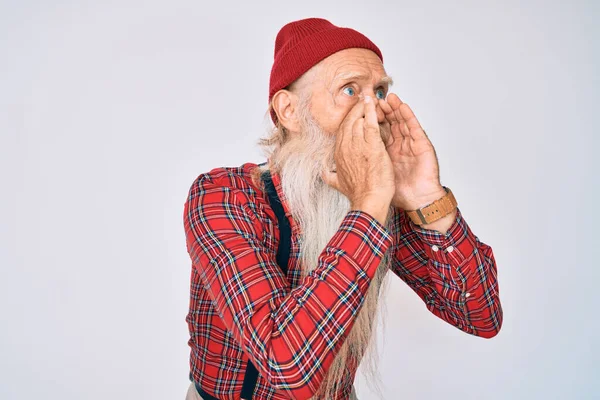 Old senior man with grey hair and long beard wearing hipster look with wool cap shouting angry out loud with hands over mouth