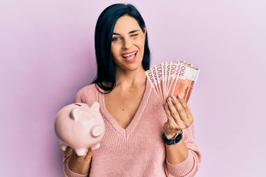 Norveç kronu banknotları tutan beyaz bir kadın ve kumbarası kameraya seksi bir ifadeyle bakıyor, neşeli ve mutlu bir yüzle.. 