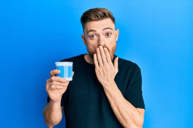 Young redhead man holding earwax cotton remover covering mouth with hand, shocked and afraid for mistake. surprised expression 