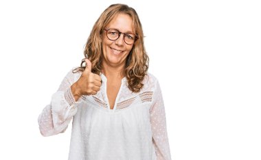 Orta yaşlı, beyaz gömlek ve gözlük takan sarışın bir kadın mutlu bir el hareketi yapıyor. Kameraya bakıp başarıyı gösteren ifade. 