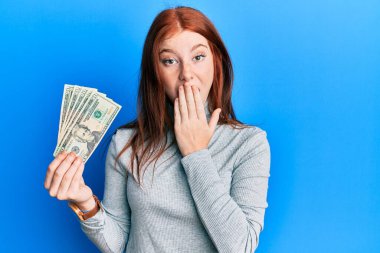 Genç kızıl saçlı kız elinde 20 dolarlık banknotlarla ağzı elle kapatıyor, şaşırmış ve hata yapmaktan korkuyor. şaşırmış ifade 
