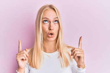 Gündelik kıyafetler giyen genç sarışın kız şaşırmış ve şaşırmış bir şekilde yukarı bakıyor ve parmaklarıyla işaret ediyor ve kollarını kaldırıyor.. 